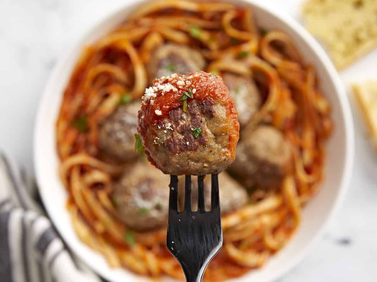 Close up of a turkey meatball on a fork with a bowl of pasta in the background.
