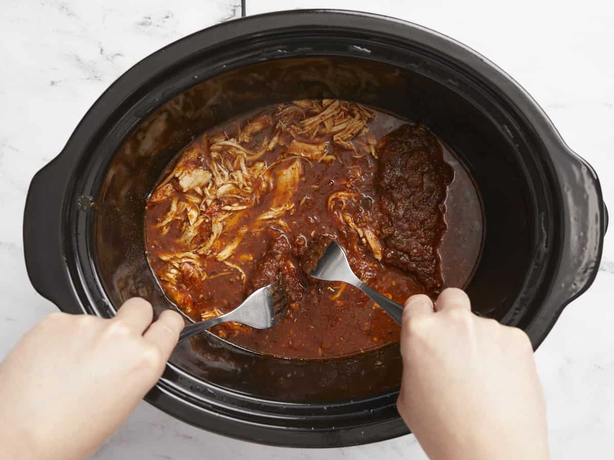 Chicken being shredded in the slow cooker with two forks.