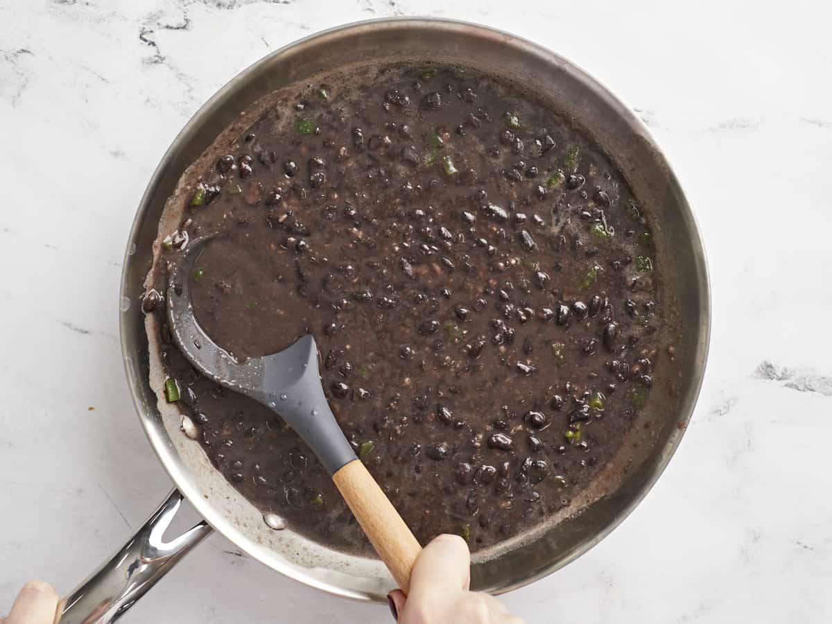 Simmered black beans in the skillet being smashed with the back of a spoon.