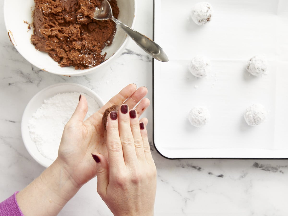 Chocolate crinkle cookies being shaped and rolled in powdered sugar.
