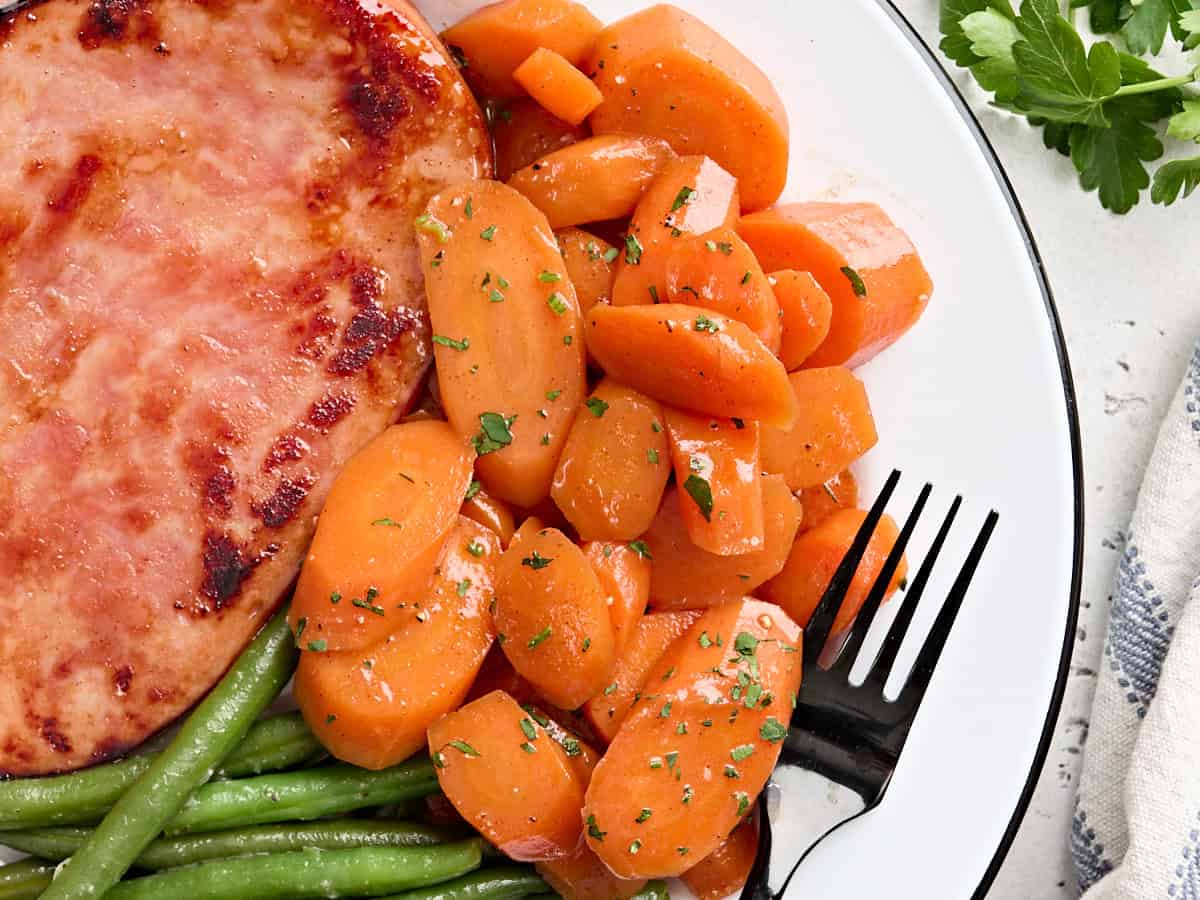 Overhead view of glazed carrots on a plate with green beans and a ham steak.