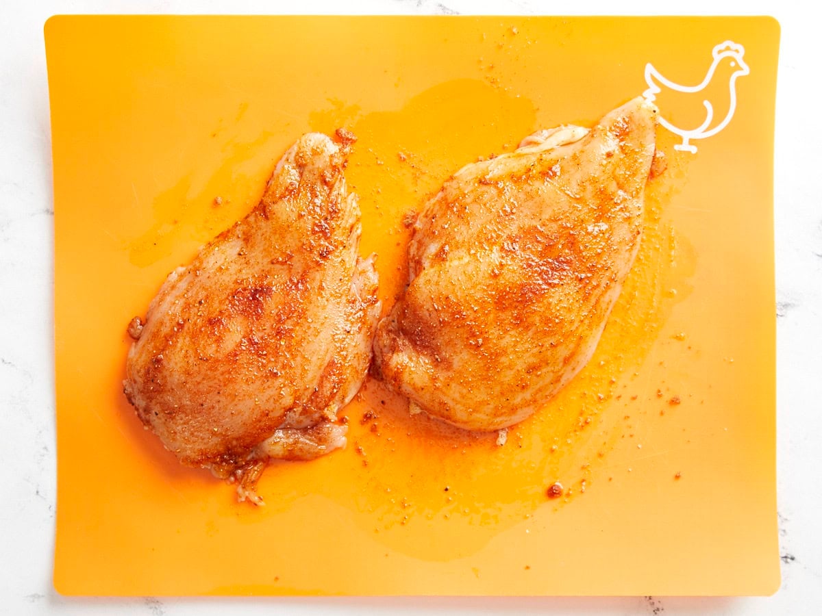 Seasoned raw chicken breasts on an orange cutting board.