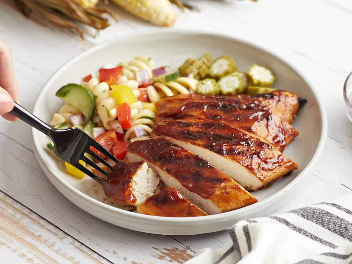 A serving of sliced grilled bbq chicken on a white plate with pasta salad and okra.