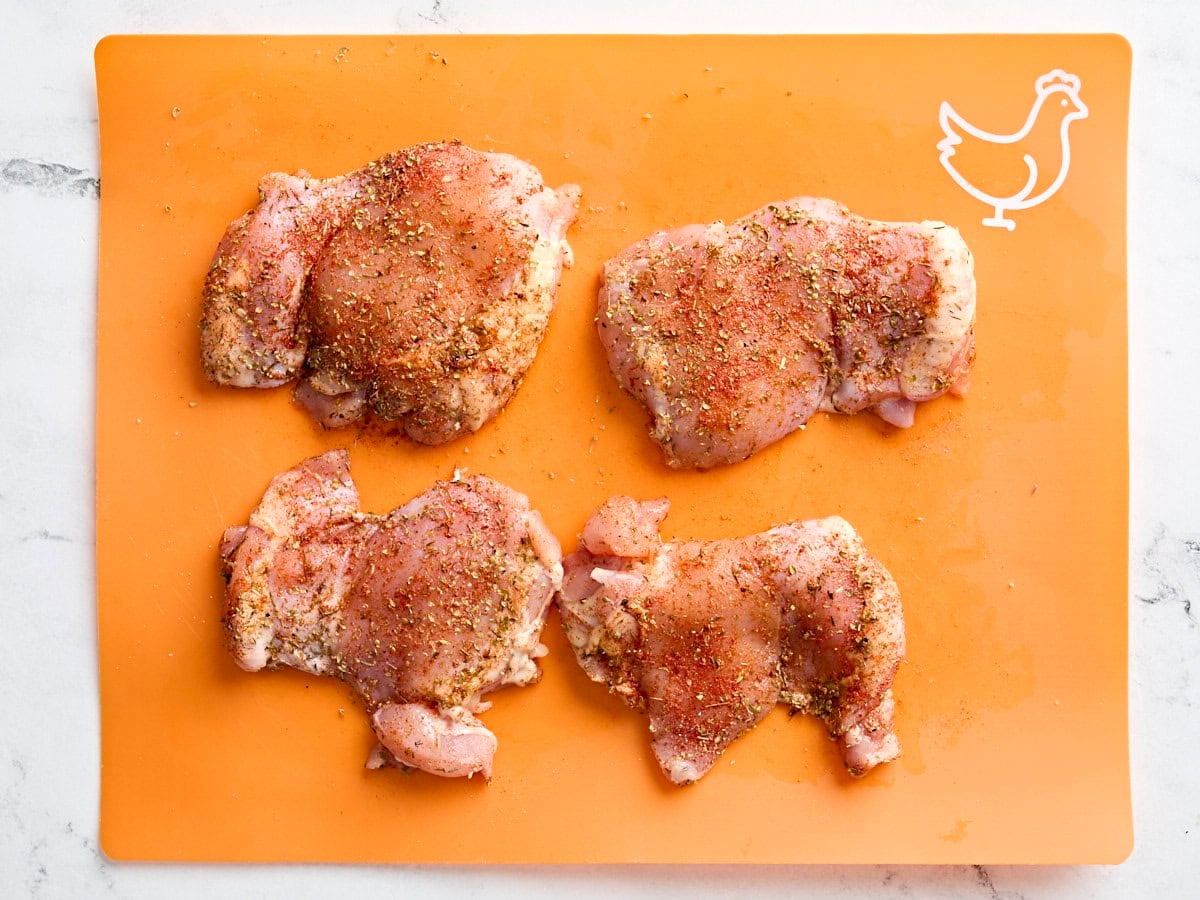 Chicken thighs seasoned with a Greek seasoning mix on a meat preparation mat.