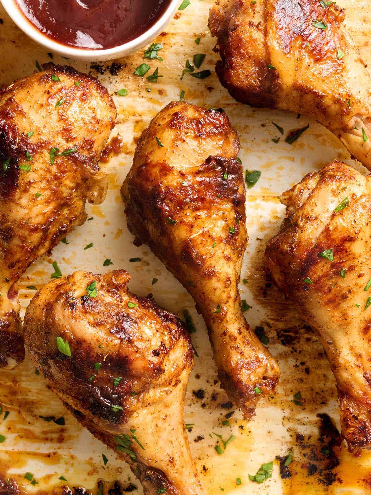 Overhead view of baked chicken drumsticks on a baking sheet with a pot of BBQ sauce.