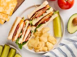 Overhead view of a chicken club sandwich on a plate with chips and a pickle.