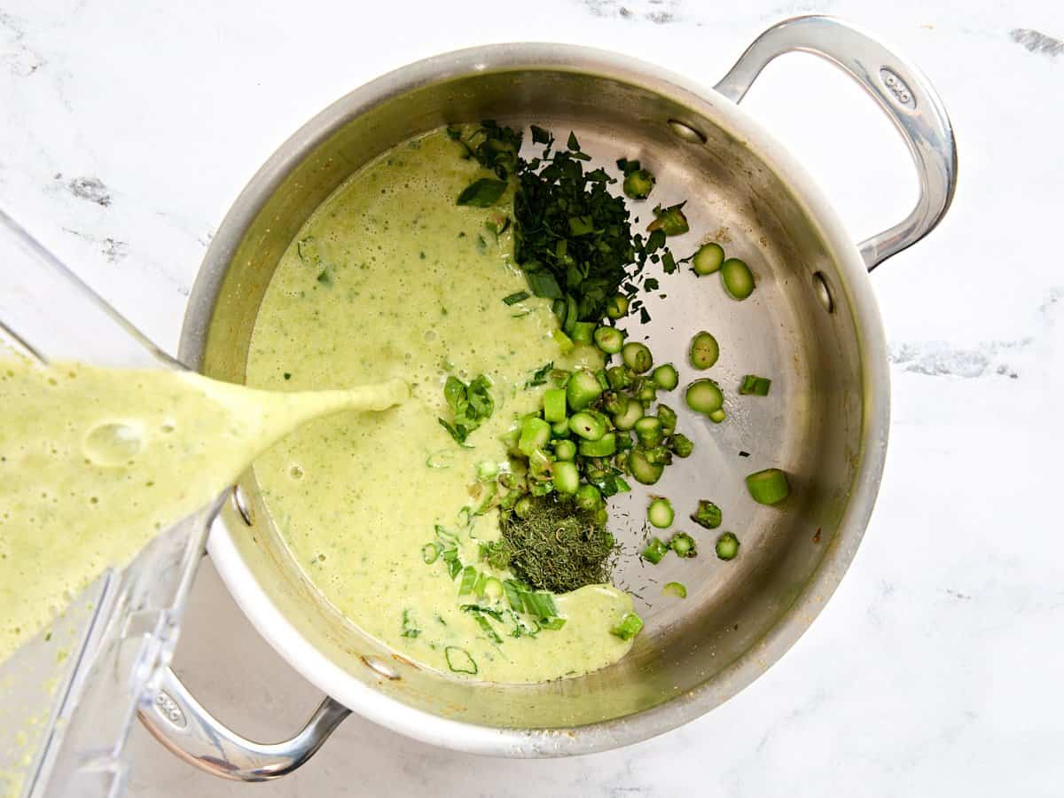 Blended homemade asparagus soup being added to a sauce pot with sliced asparagus, green onion, and parsley.