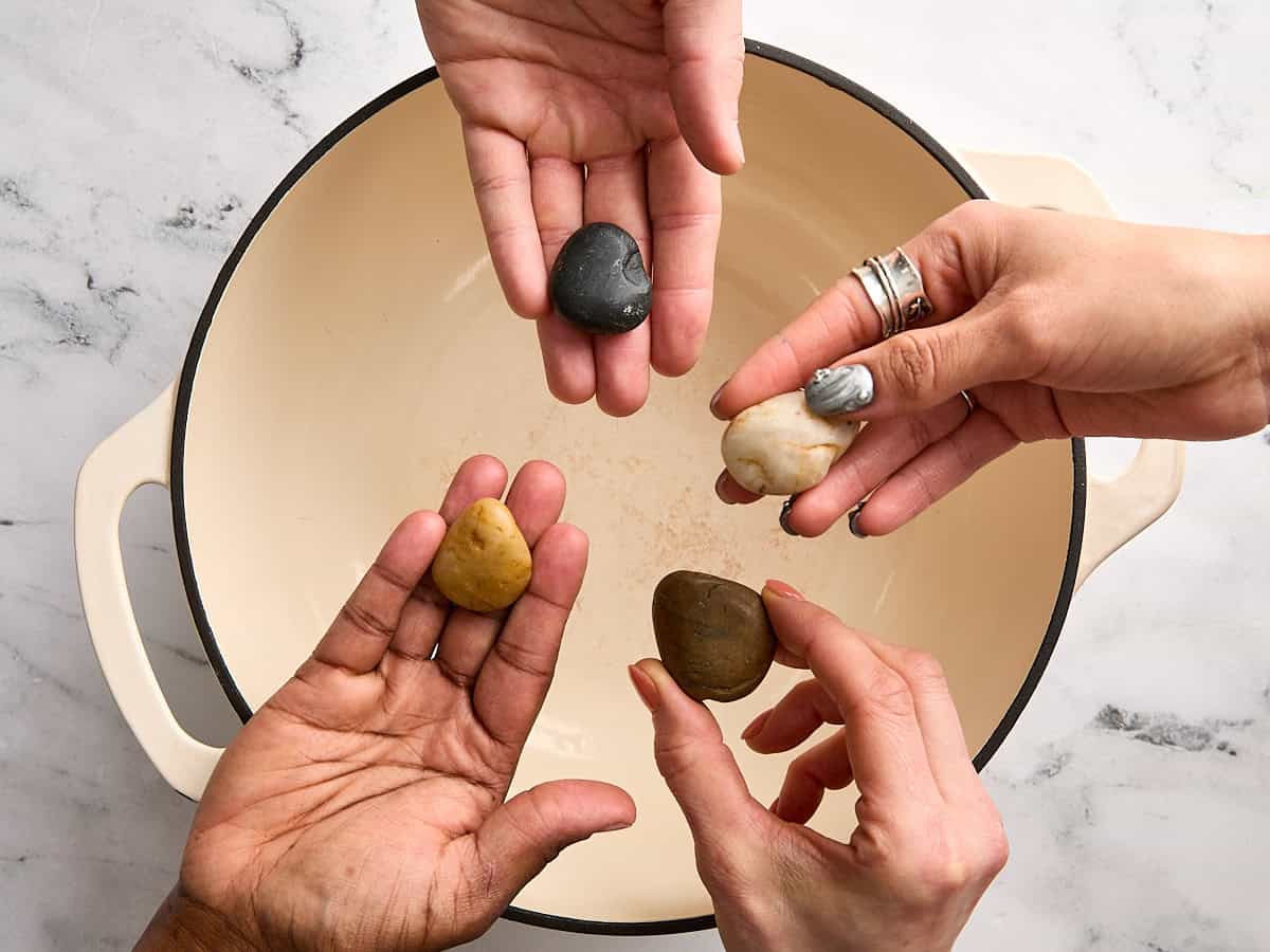 Four hands holding a small river rock over a stock pot.