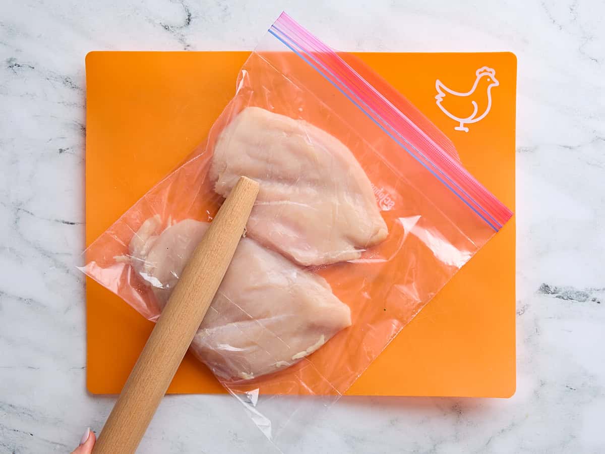 Chicken breasts in a ziploc bag, being pounded with a wooden rolling pin.