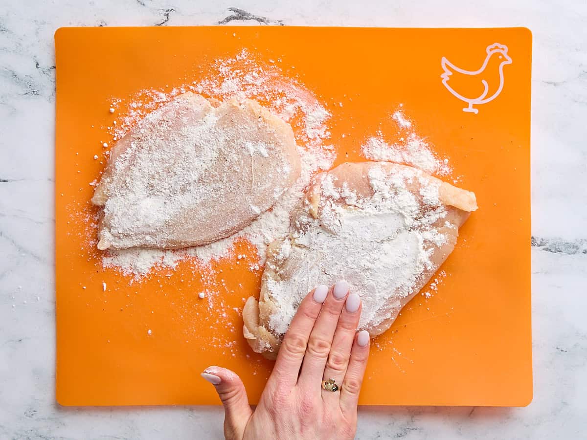 A hand coating two pounded chicken breasts with flour.