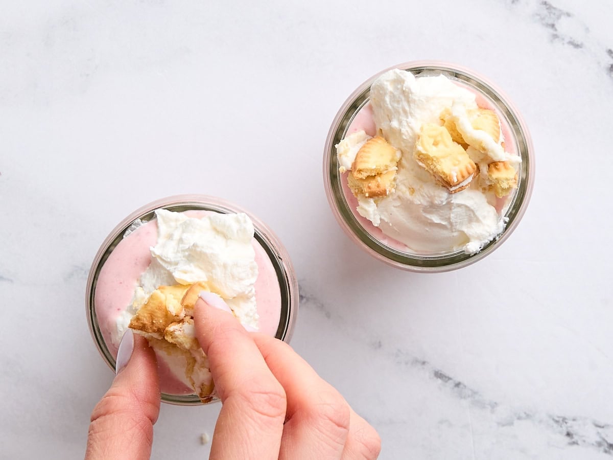 Two glasses of strawberry cheesecake smoothie, topped with whipped cream and crumbled vanilla sandwich cookies.