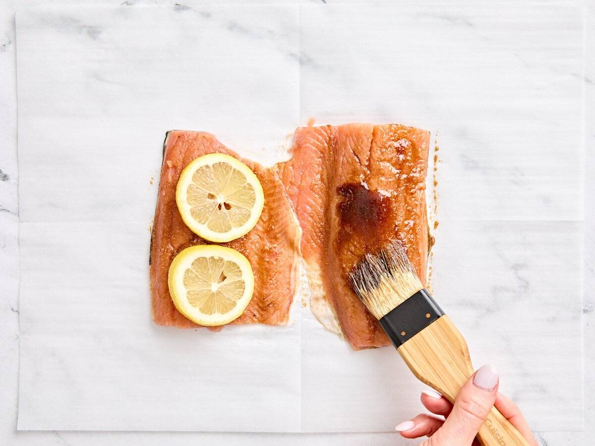 A brush glazing two salmon filets with a toasted sesame oil glaze. One filet is topped with two lemon slices.