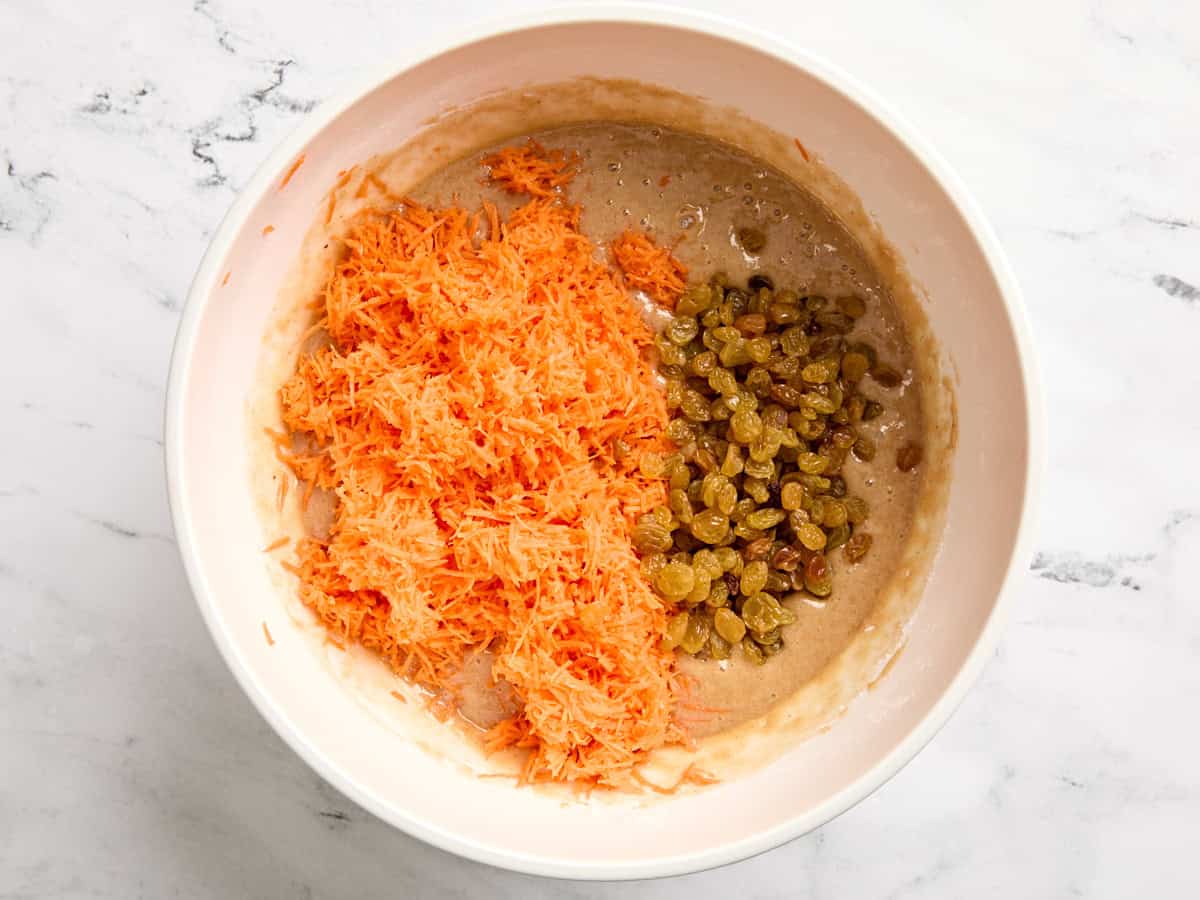 Shredded carrots and golden raisins added to cake batter in a mixing bowl.