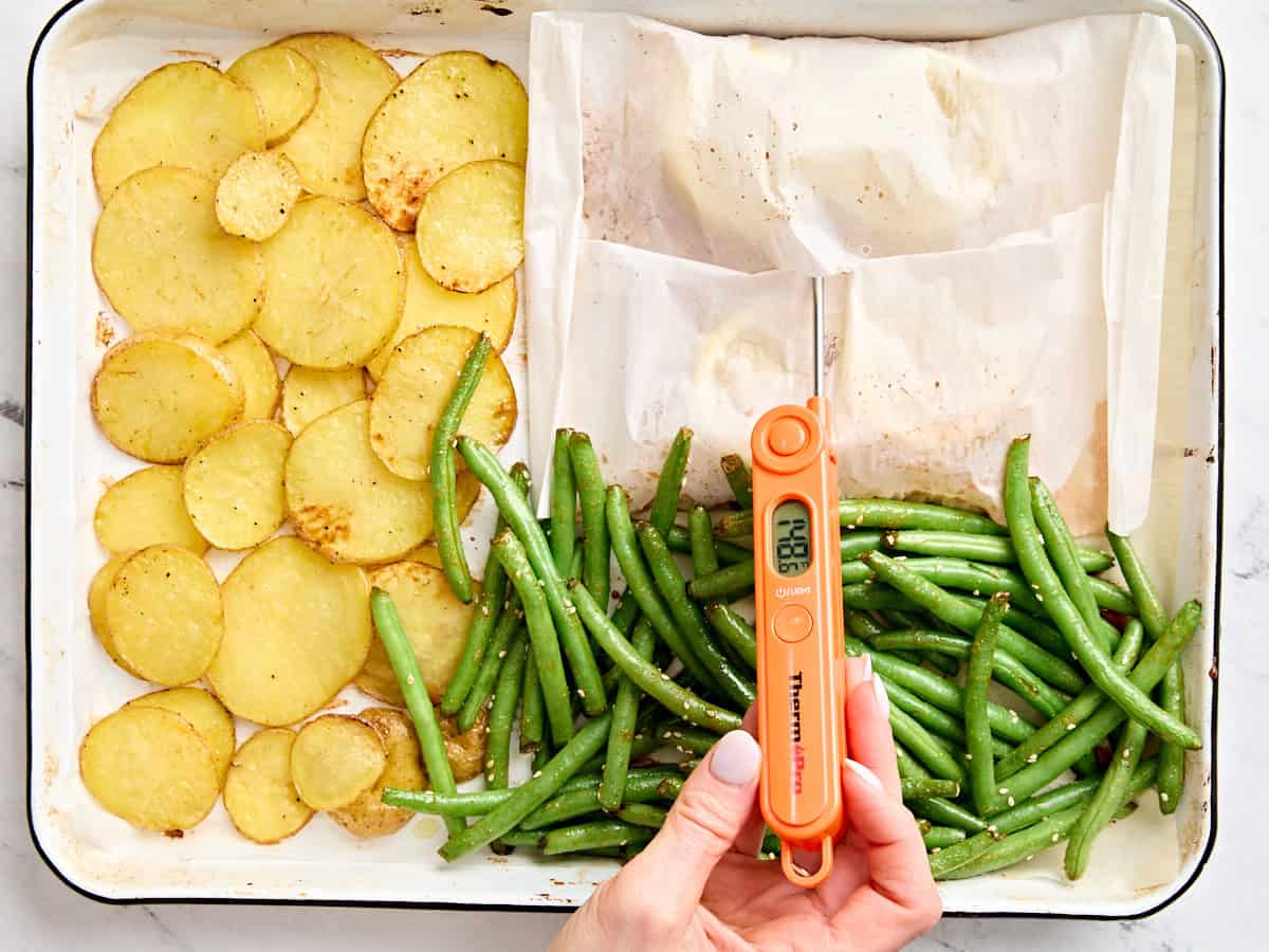 Sliced potatoes, green beans, and parchment paper wrapped salmon filets on a baking sheet, with a instant read thermometer checking the internal temperature of the salmon.