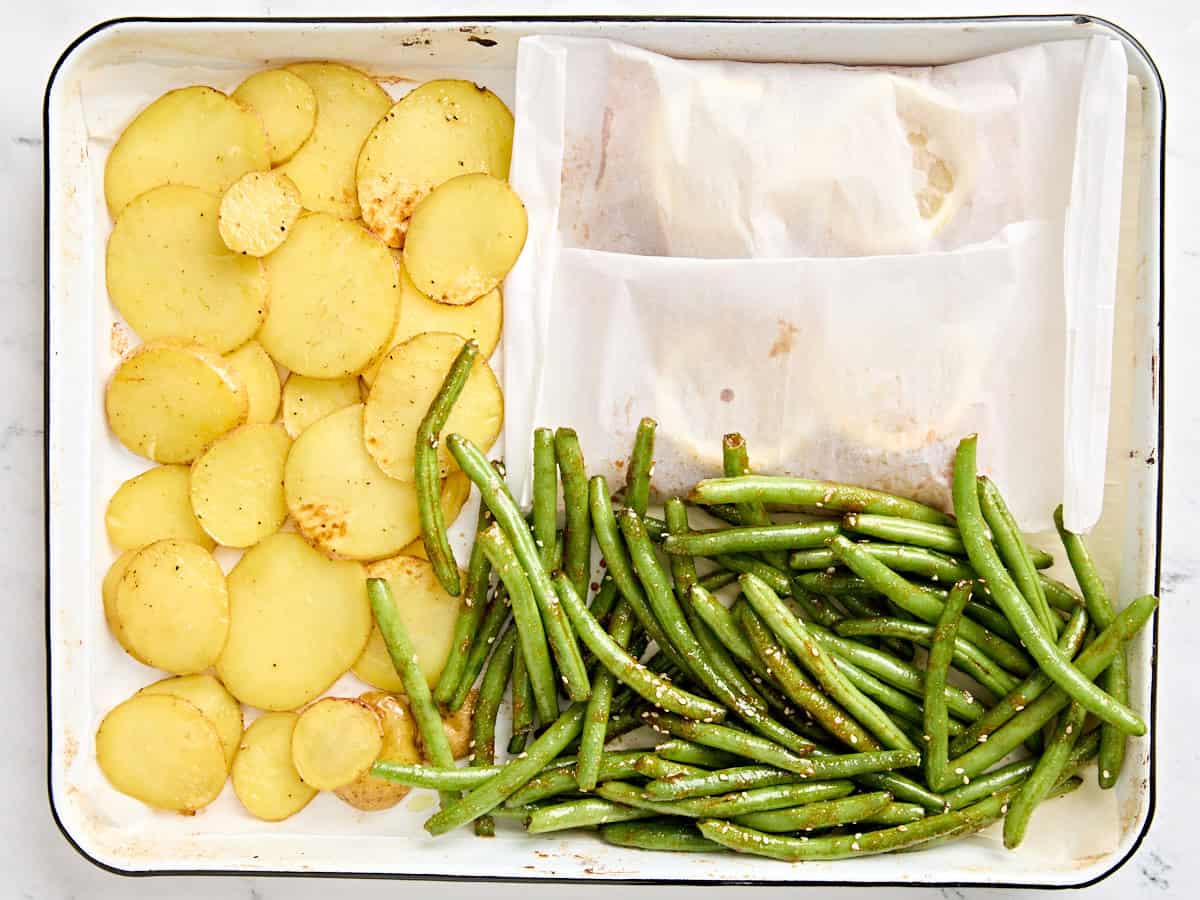 Sliced potatoes, green beans, and parchment paper wrapped salmon filets on a baking sheet.