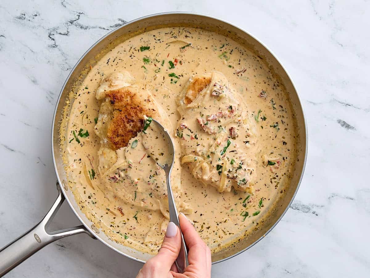 Pan fried chicken breasts added to the sauce for marry me chicken in a skillet, with a spoon pouring the sauce overtop.