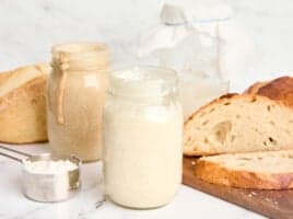 Side view of two jars of sourdough starter with a loaf of sourdough bread.