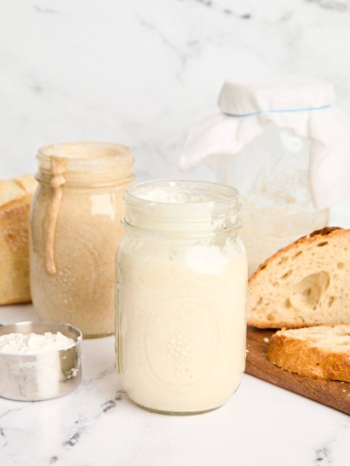 Side view of two jars of sourdough starter with a loaf of sourdough bread.