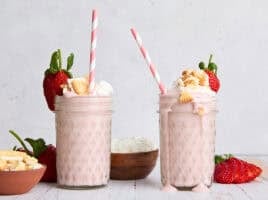 Side view of two strawberry cheesecake smoothies, topped with whipped cream, crumbled cookies, and strawberries.