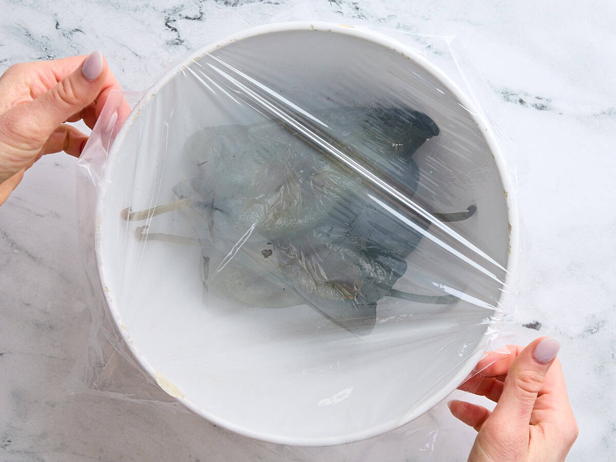 Roasted poblano peppers in a bowl, with hands covering them with plastic wrap to steam.