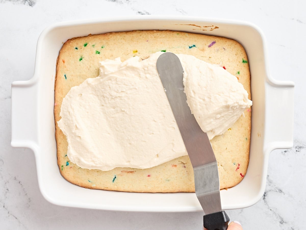 Vanilla frosting being spread onto a sugar cookie bar base in a baking dish.