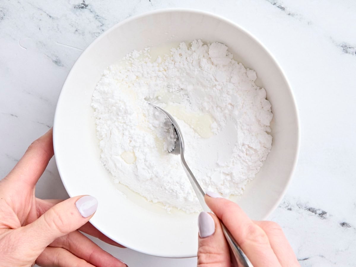 Powdered sugar and milk in a mixing bowl, being mixed with a spoon to make a glaze.