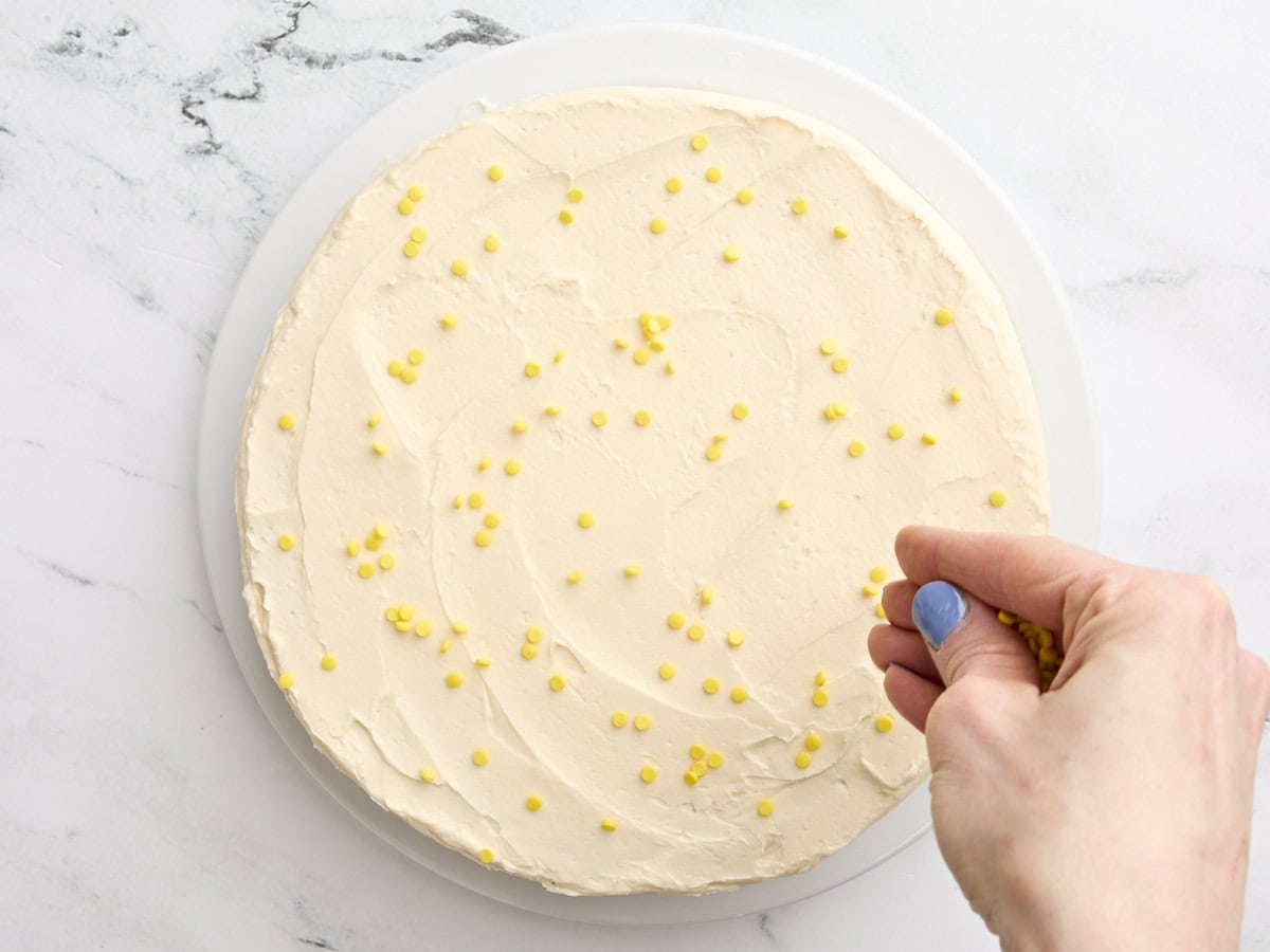 A hand sprinkling yellow sprinkles over a finished cornbread cake with honey buttercream.