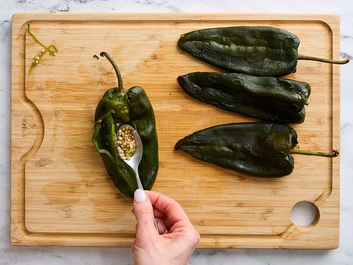 A hand holding a spoon scooping out the seeds from a roasted poblano pepper.