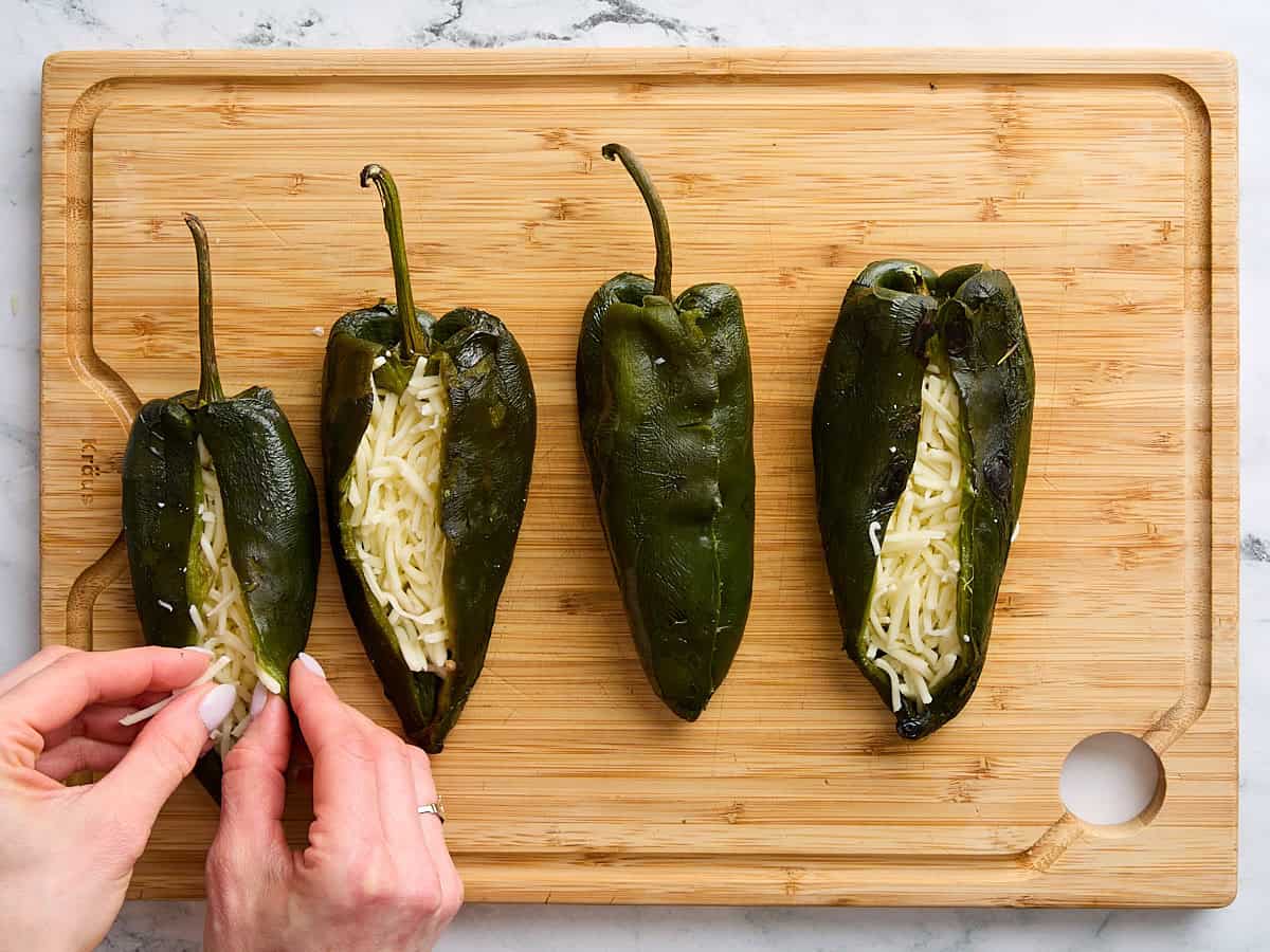 Roasted poblano peppers on a wooden cutting board being stuffed with shredded cheese.
