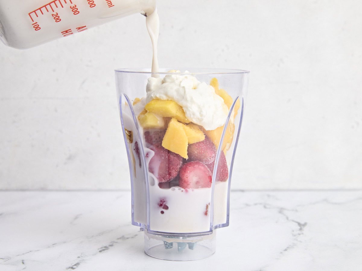 The ingredients to make a strawberry mango smoothie in a blender, with the almond milk being poured in.