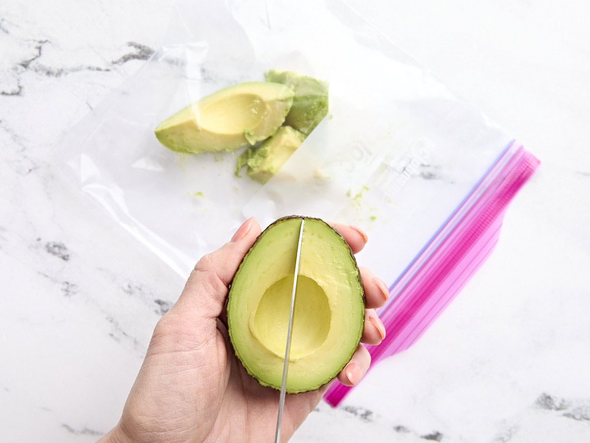 A hand holding half an avocado with a knife slicing the flesh.