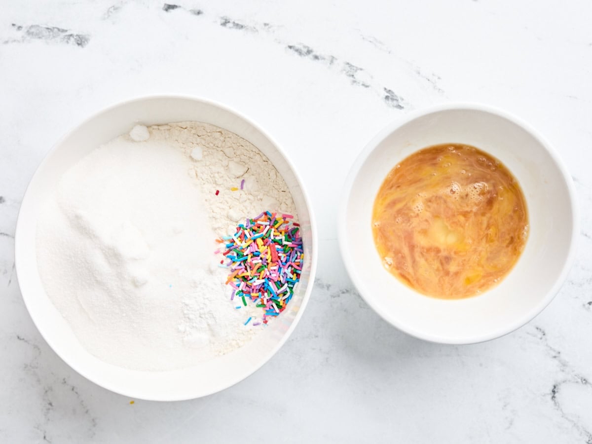 Overhead view of two mixing bowls, one with flour, sugar and sprinkles, and the other with eggs and vanilla.