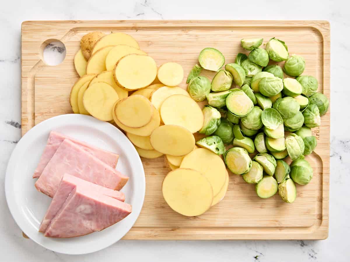 Sliced ham steak, potatoes, and Brussels sprouts on a wooden cutting board.