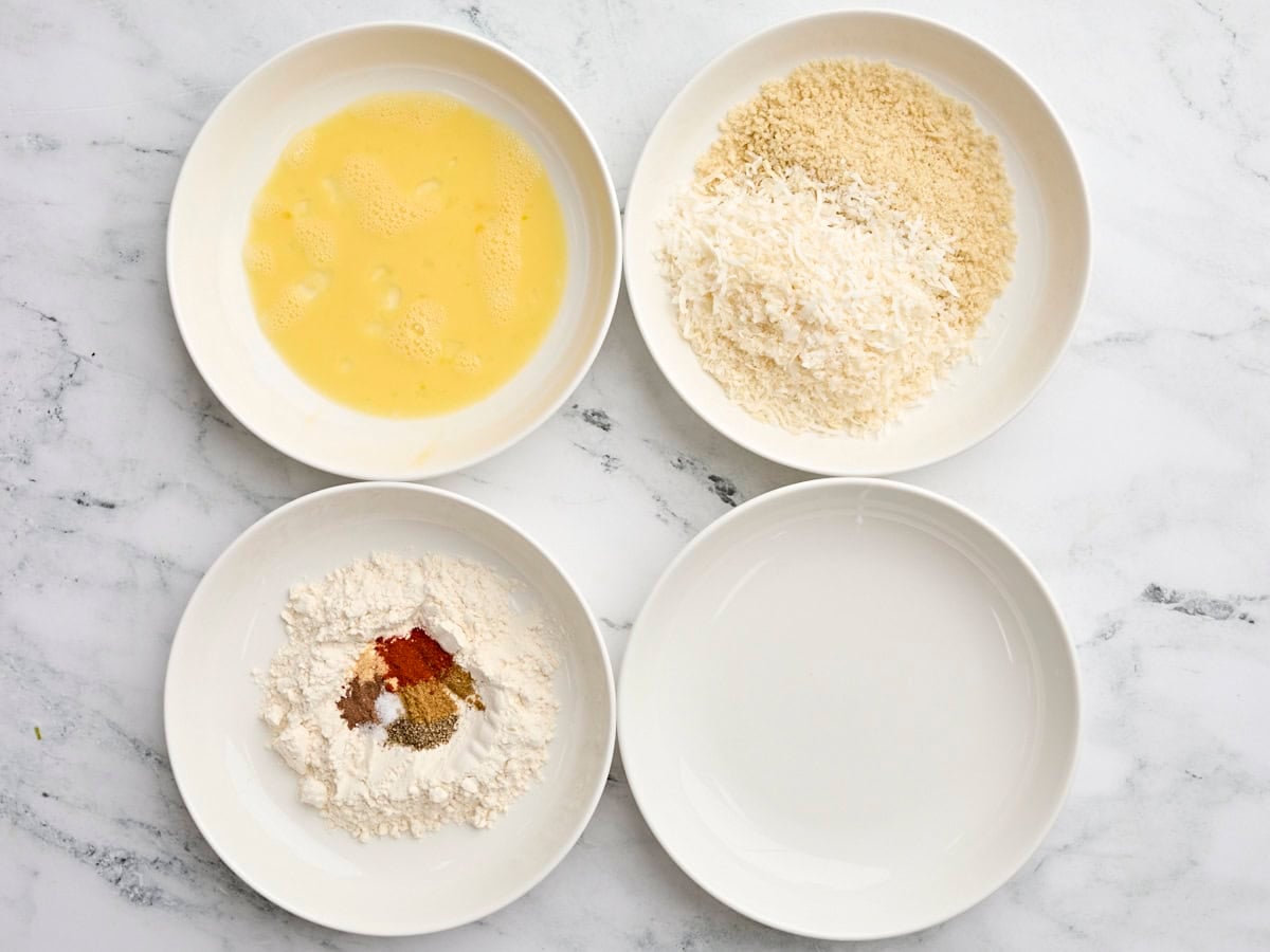 A bowl of egg wash, a bowl of shredded coconut and panko, a bowl of flour and seasonings, and an empty bowl.