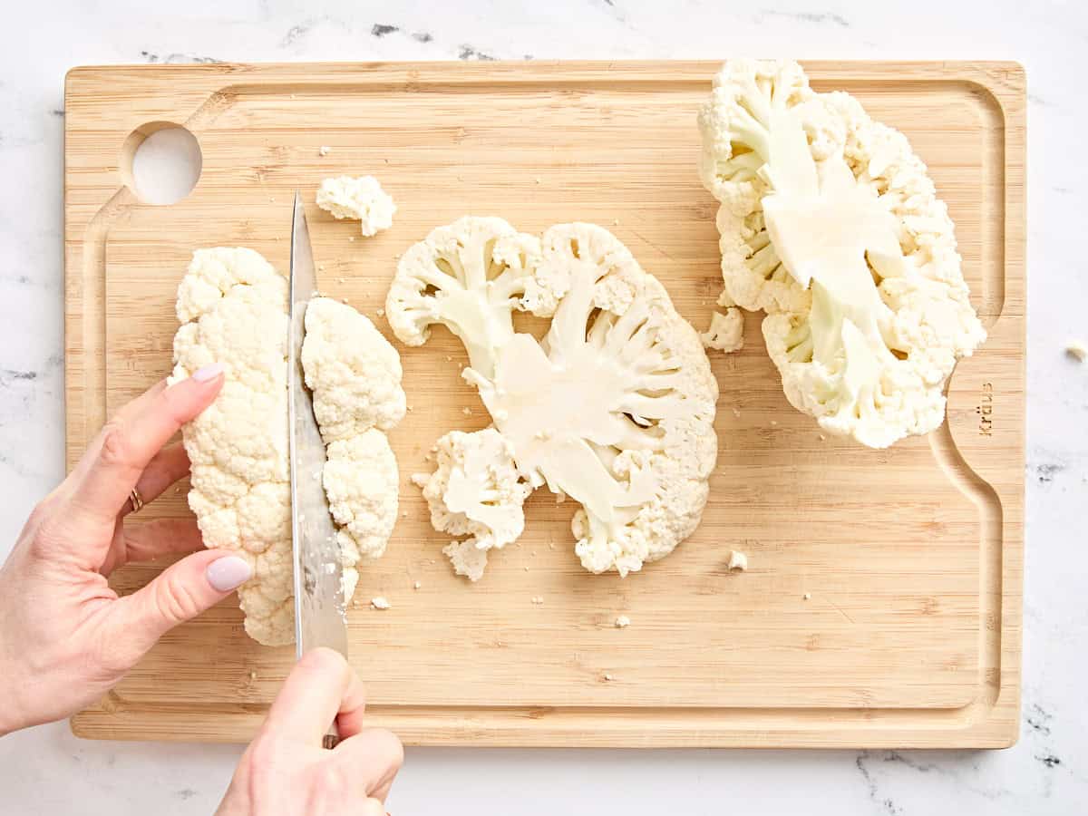 A knife slicing a head of cauliflower into steaks on a cutting board.