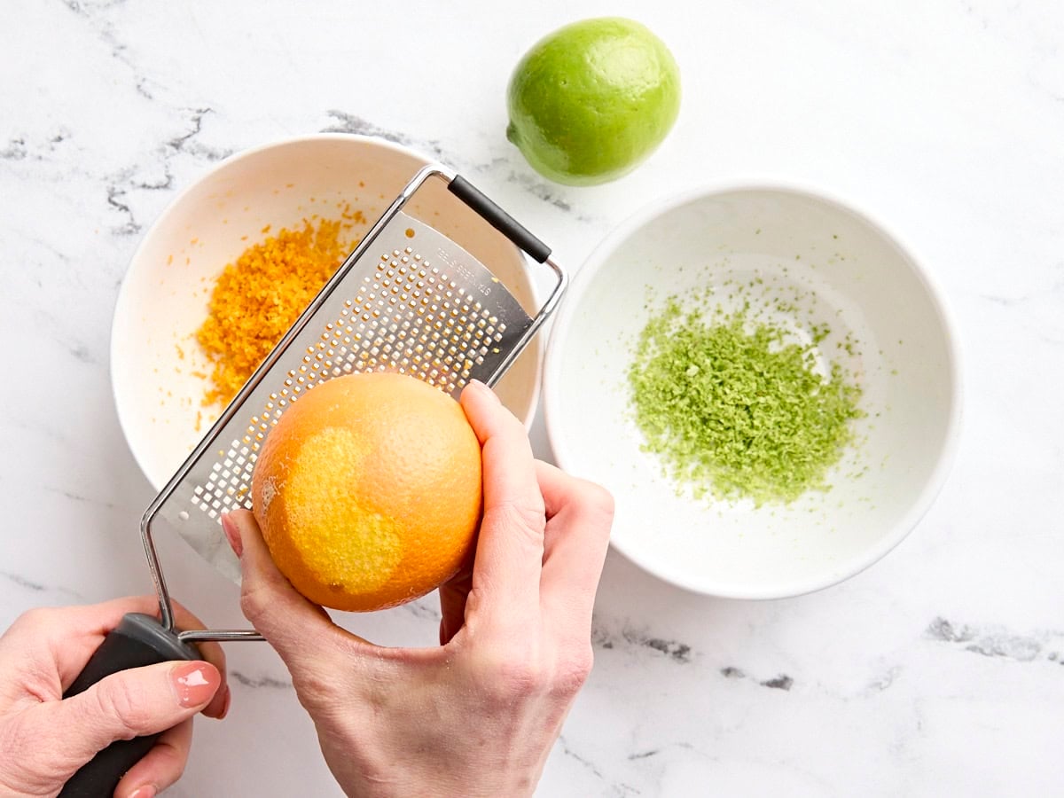 A bowl of lime zest and a hand zesting an orange over another bowl.