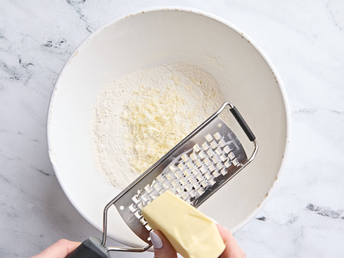 Butter being grated into a mixing bowl of flour, baking powder, sugar, and salt.