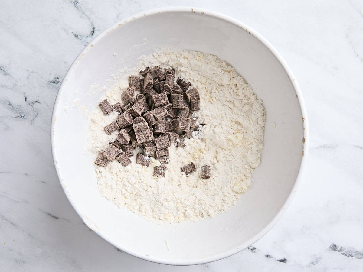 Chocolate chunks added to a four and butter mixture in a white mixing bowl.