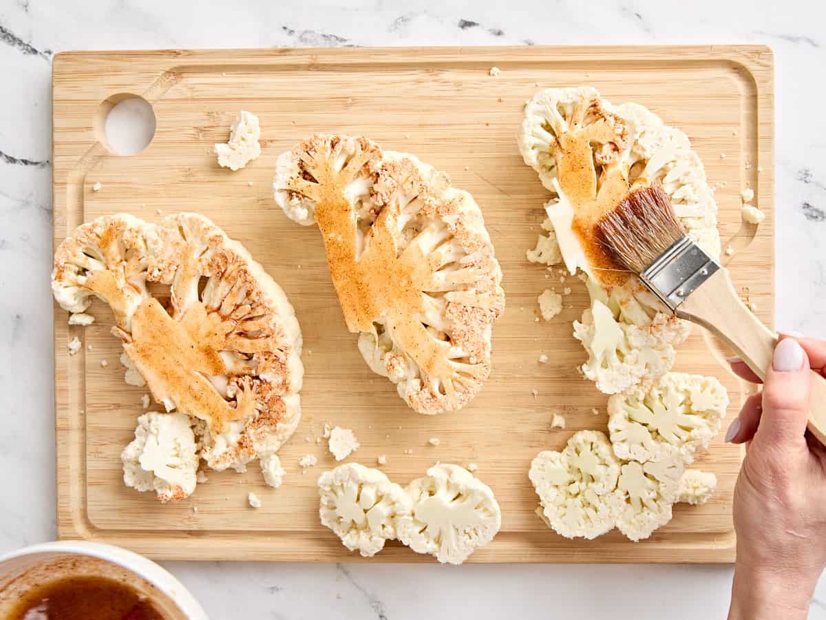 A brush adding a seasoning marinade to cauliflower steaks on a wooden cutting board.