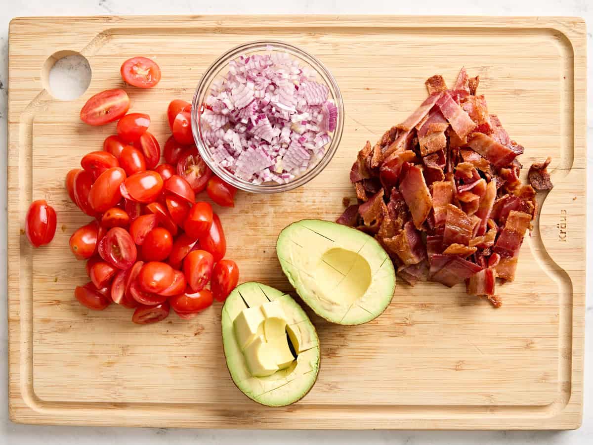 Diced avocado, halved plum tomatoes, finely diced onion, and chopped cooked bacon on a wooden chopping board.
