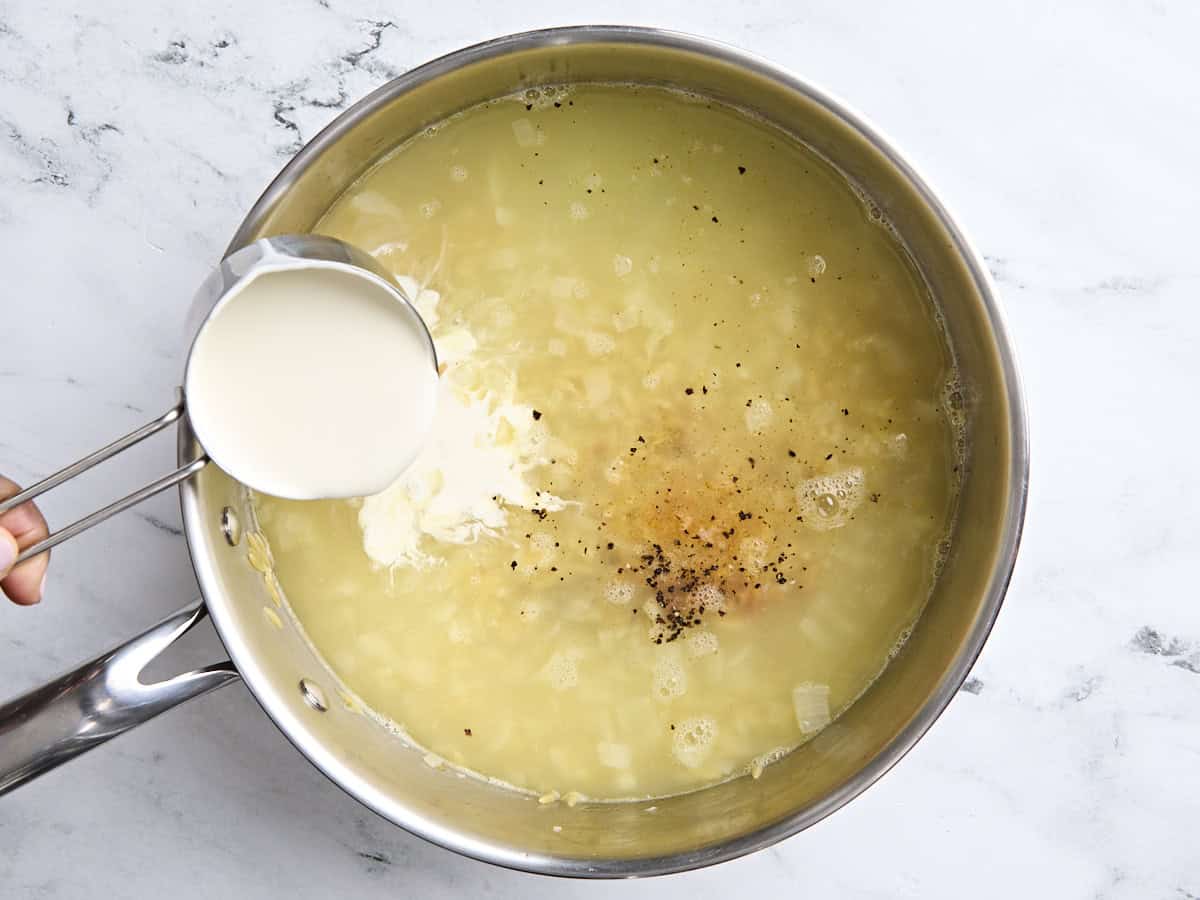 Chicken broth in a skillet with orzo, with heavy cream being poured in.