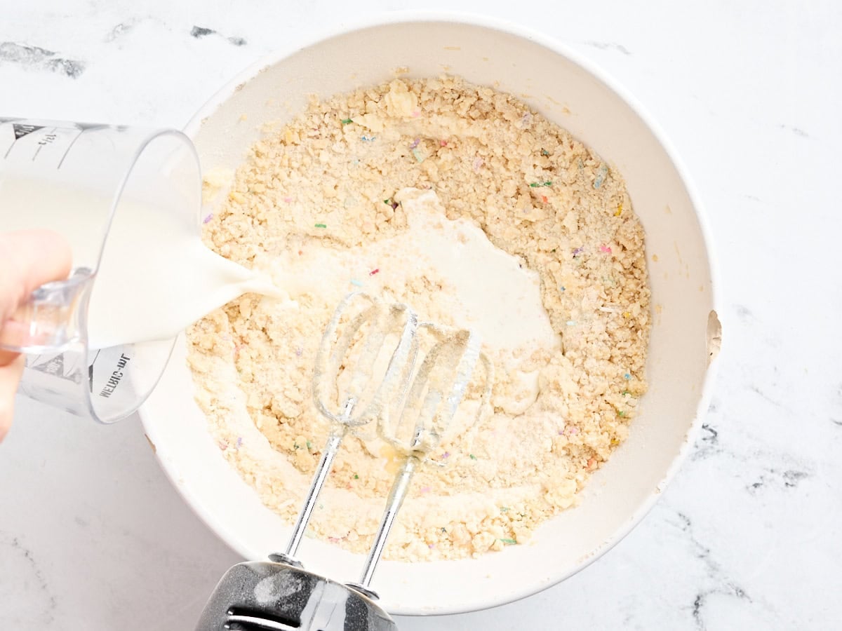Milk being poured into a flour, butter, and egg mixture in a mixing bowl.