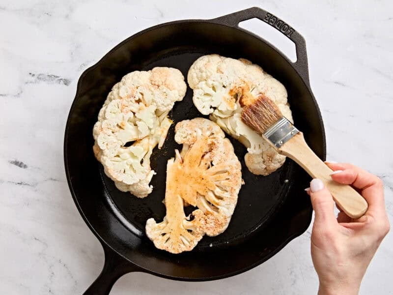 Cauliflower cooking in a skillet with more marinade being brushed over them.
