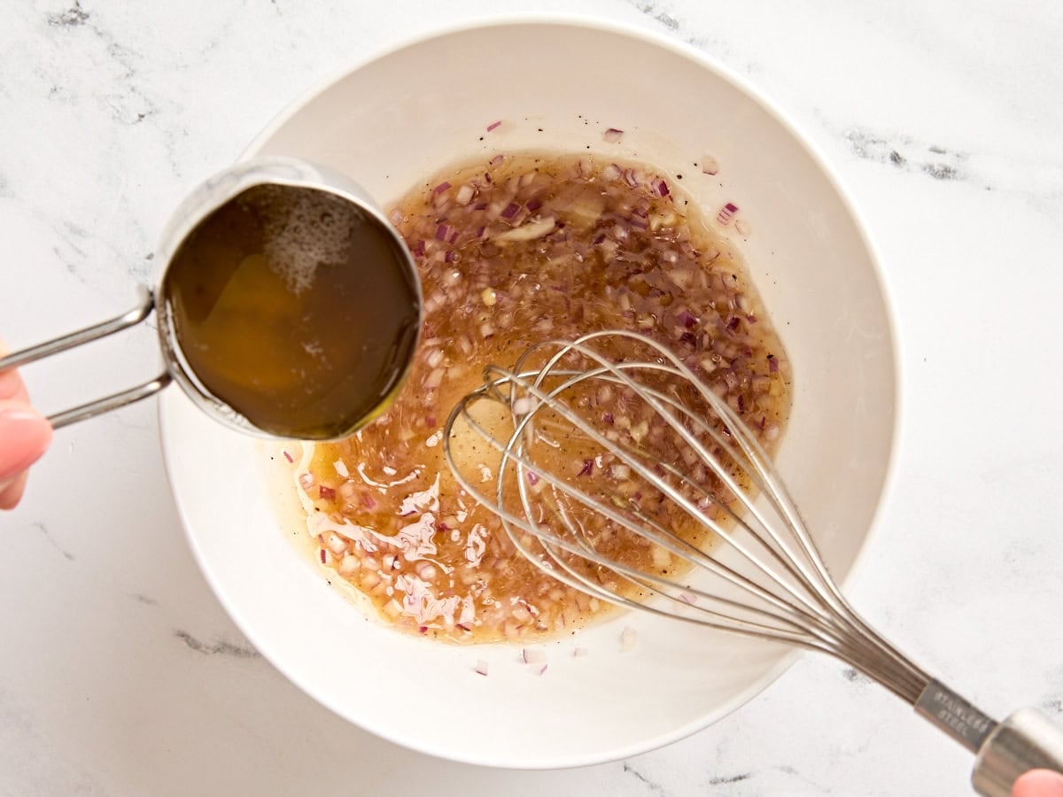 Bacon fat being poured into a bowl with minced red onion and seasonings to make a warm bacon vinaigrette.