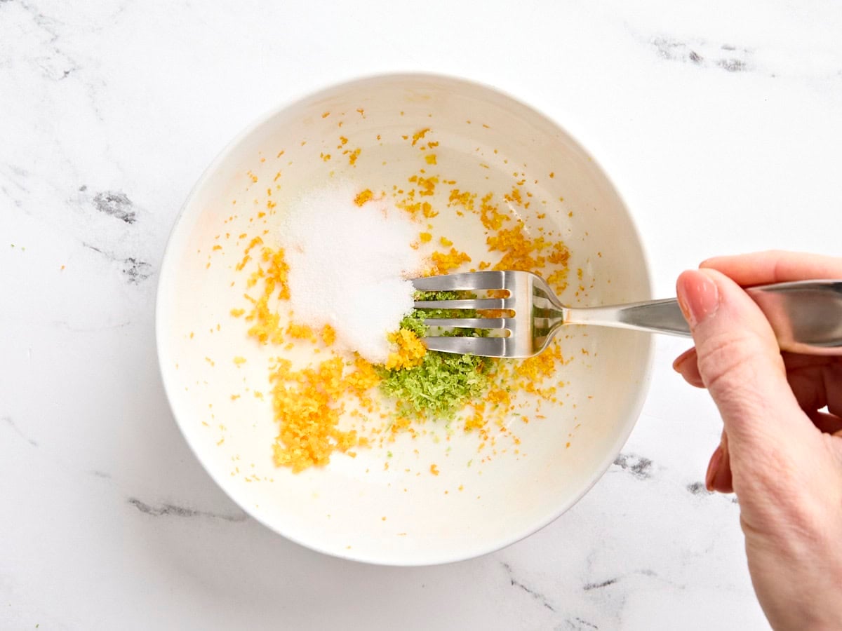 Salt, orange zest, and lime zest being mixed with a fork in a bowl.