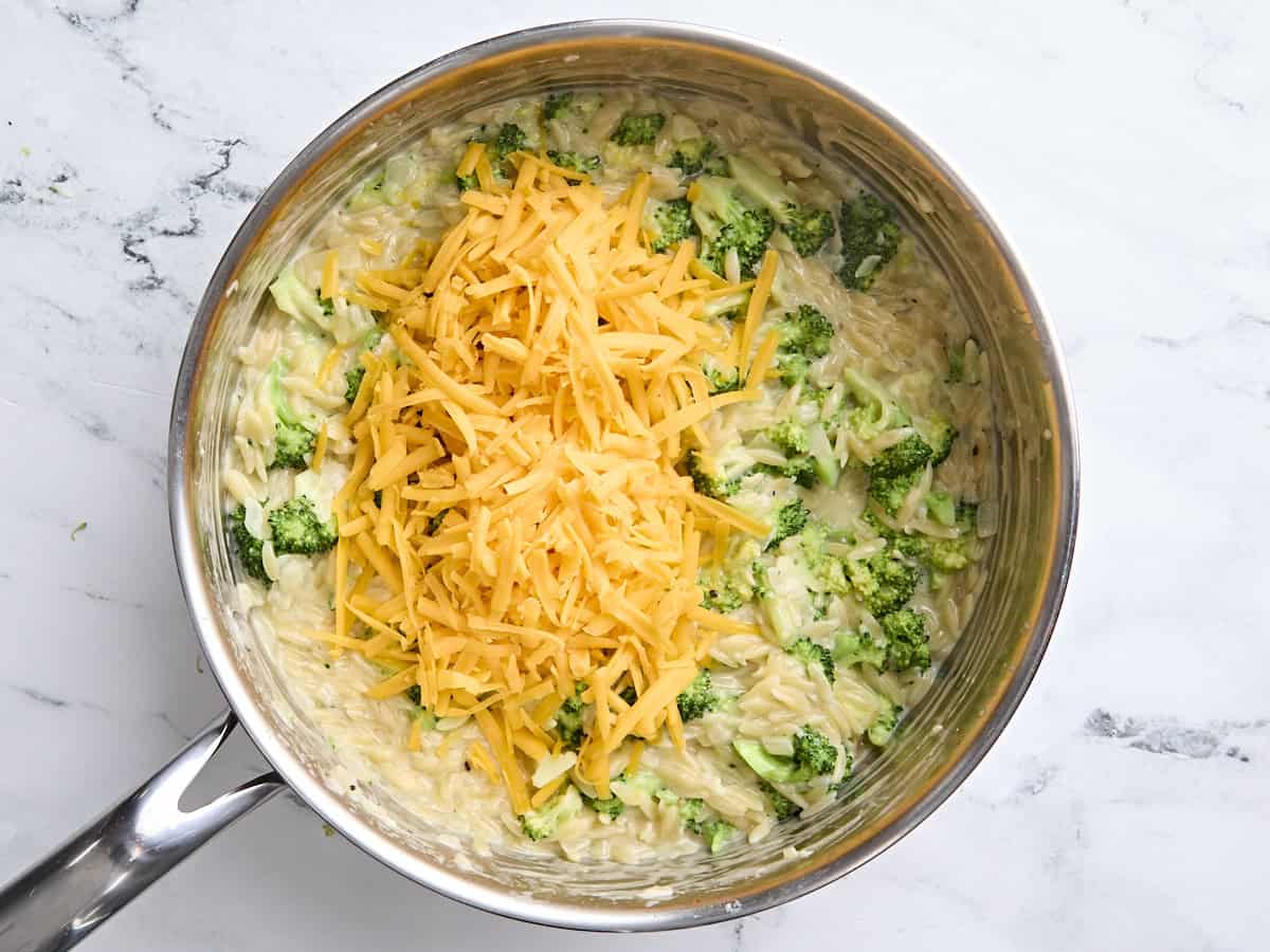Shredded cheese added to the broccoli cheese orzo in a skillet.