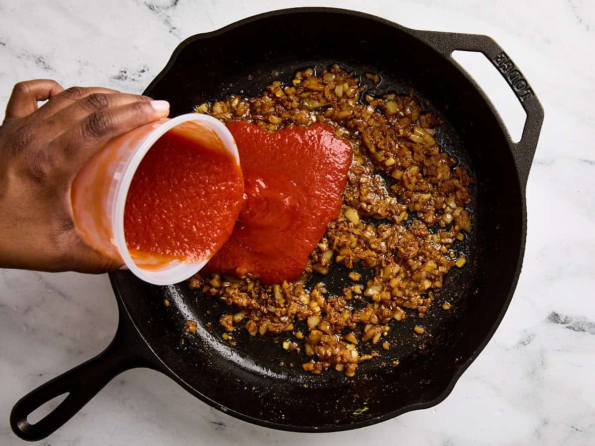 Tomato sauce being poured into spiced sautéed onions in a skillet.