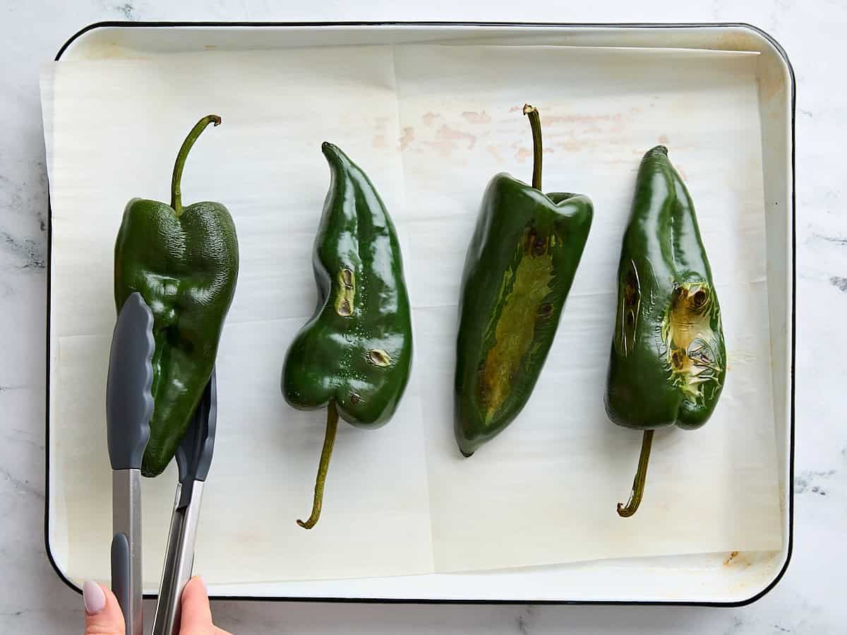 Kitchen tongs flipping over a roasted poblano pepper on a parchment lined baking sheet.