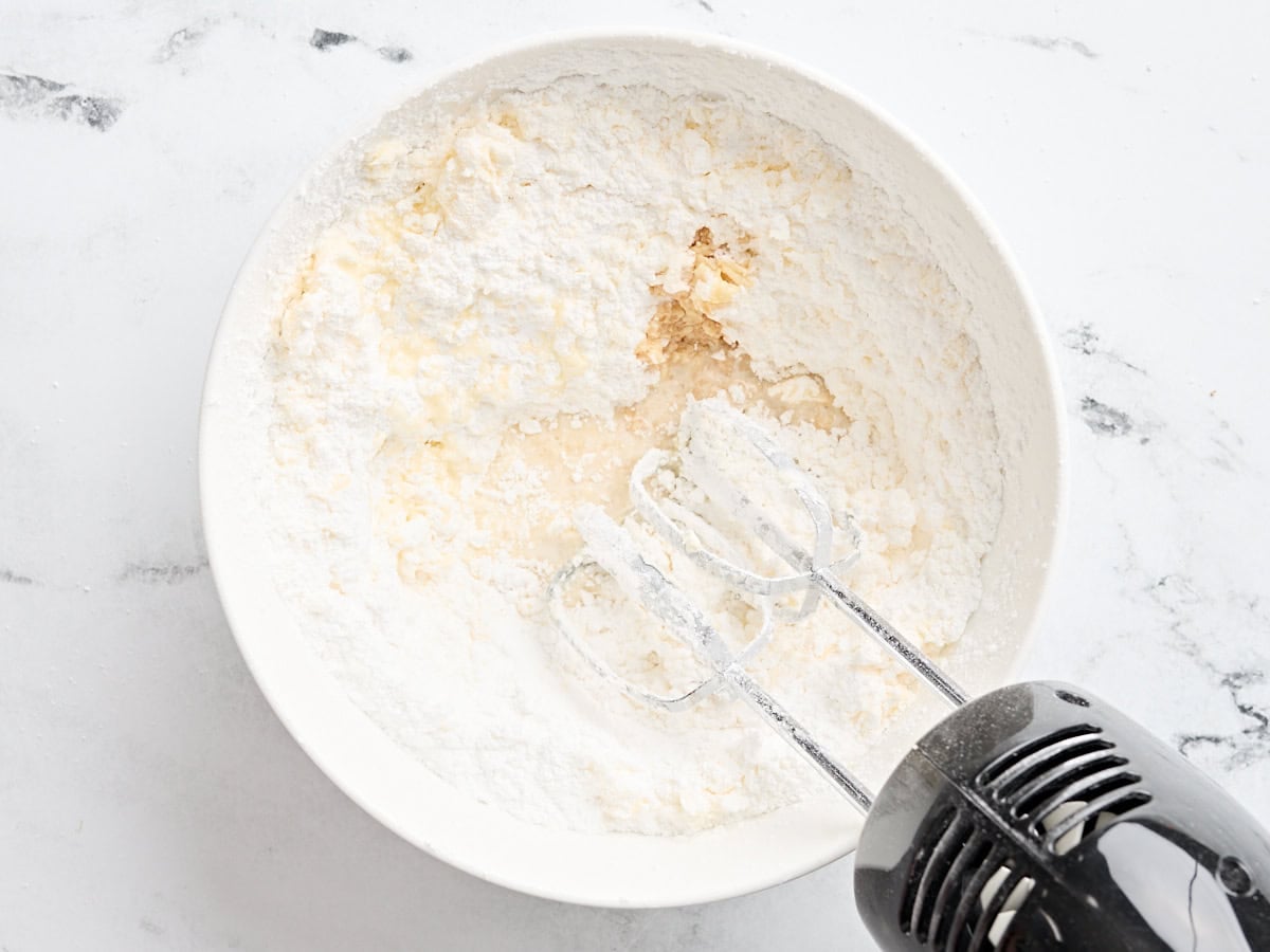 Powdered sugar, butter, and vanilla being mixed in a mixing bowl to make frosting.