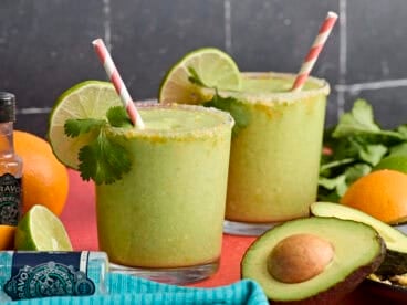Side view of two glasses of avocado margarita, garnished with lime slices and fresh cilantro.